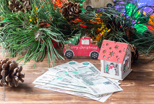 Russian ruble banknotes next to a toy house and a red toy truck on a wooden table with pine branches.