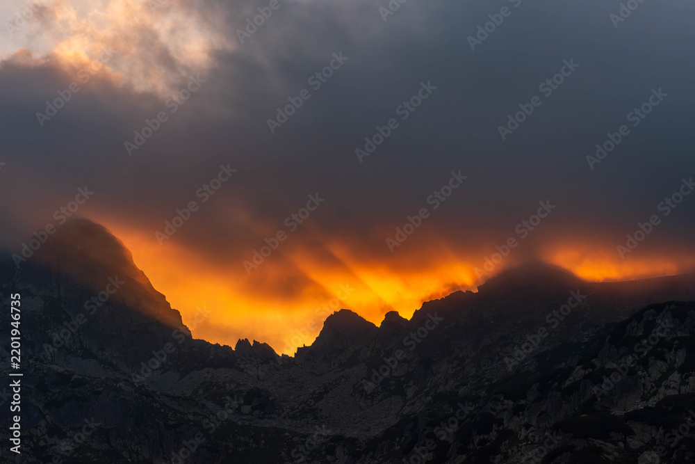 Obraz premium Dramatic Golden Sunbeams Piercing Storm Clouds Over Mountain Peaks at Sunset