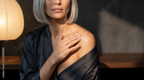 Mature woman applying moisturizer cream to her bare shoulder skin closeup