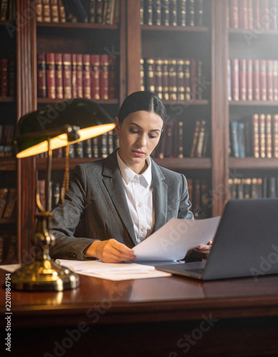 A professional female lawyer studies legal documents in a dimly lit, atmospheric library office. This elegant scene evokes focus, authority, and scholarly dedication to the practice of law.