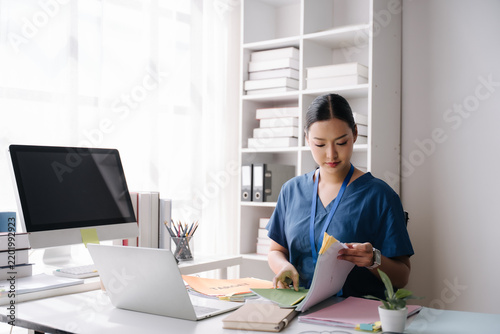 Fotografie Focused Professional at Work: A focused female professional, likely a doctor or nurse given her scrubs, meticulously organizes medical documents at a well-lit desk with a computer and laptop