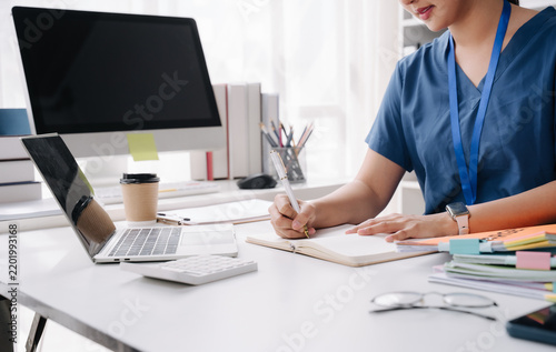 Doctor studying in the office: A doctor in a blue scrub suit meticulously takes notes in a notebook while working on a laptop and computer at a desk.