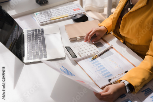 Professional Analysis: A woman in a vibrant yellow blazer meticulously reviews financial reports and performs calculations at her desk, illuminated by natural light.