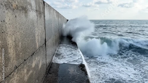 Massive ocean wave violently crashes against a sturdy concrete seawall, illustrating the raw power and untamed force of nature meeting human-made coastal defenses under a dramatic sky