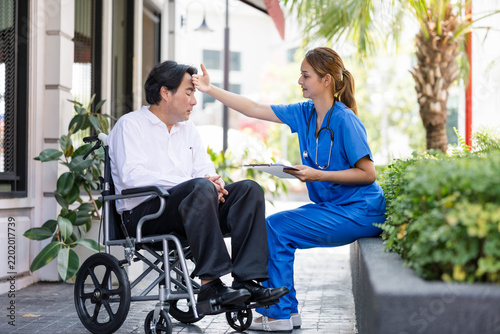 Caregiver in scrubs checking a man in a wheelchair outdoors, offering support and medical attention on a city sidewalk with plants and palm trees.