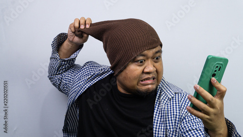 Fotografie An angry adult Asian male forcefully pulls a brown knit beanie while glaring at a green smartphone