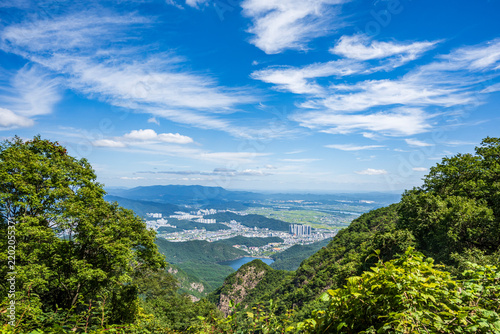 Panoramic view of lush green mountains overlooking a distant city and blue lake under a clear sky with white clouds.