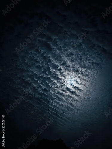Full moon shining brightly through layer of dense rippling textured clouds in dark midnight sky view.