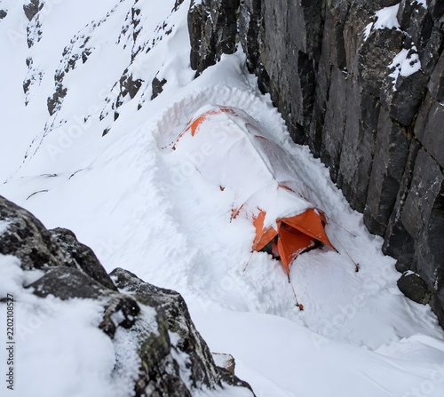 Orange mountaineering tent dug into a snow trench against a basalt cliff wall with a sagging flysheet