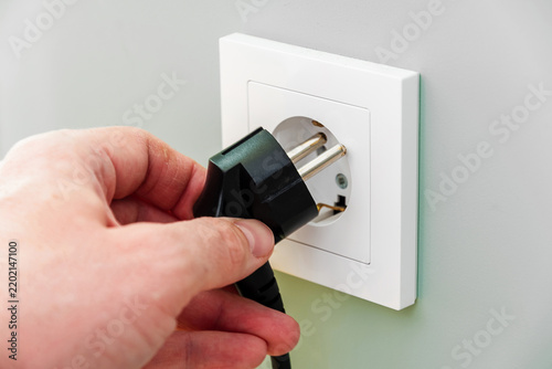 Close-up of a hand plugging a black electric cable into a white wall socket. Symbol of power connection, energy consumption, and home utilities