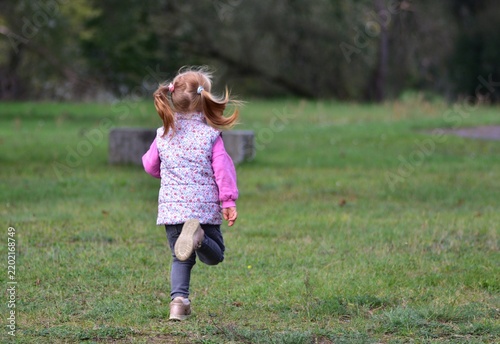 Foto Young red-haired child running away across a green meadow in a peaceful park