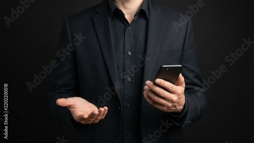 Businessman in a black suit holding a smartphone and gesturing with an open hand against a dark background, for corporate concepts