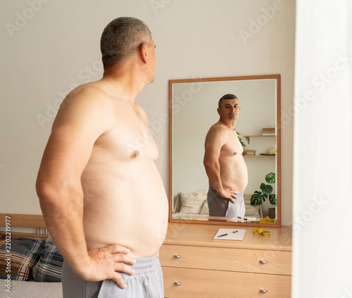 An overweight middle-aged man stands shirtless with his back to camera, looking at his reflection in mirror and assessing changes in his physique. Real person, weight and health monitoring