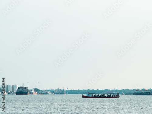 Red wooden passenger ferry boat crossing wide river with passengers, hazy harbor skyline of cargo ships and white oil storage tanks in distance, calm water with large sky copy space