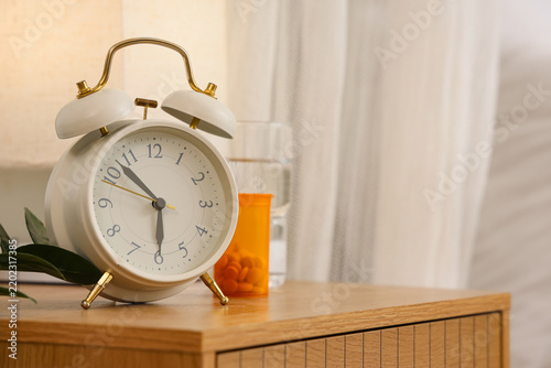 Foto Alarm clock with lamp, pill bottle and glass of water on table in room, closeup