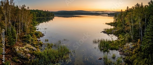 Peaceful panoramic view of a calm lake surrounded by dense forest at sunrise. Golden morning light reflects on still water in a remote wilderness landscape during early autumn.