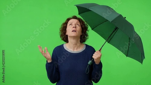 Worried woman holding an open black umbrella and looking upward against a bright green screen background, expressing confusion or concern