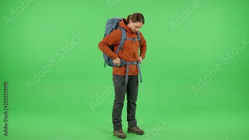 Full-body young male hiker in orange outdoor clothing and backpack checking gear against a bright green studio background