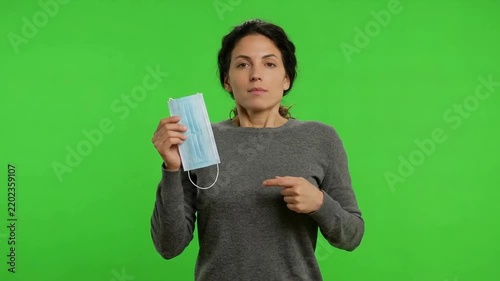Young woman holds and points to a blue disposable face mask against a bright green screen background, emphasizing health protection