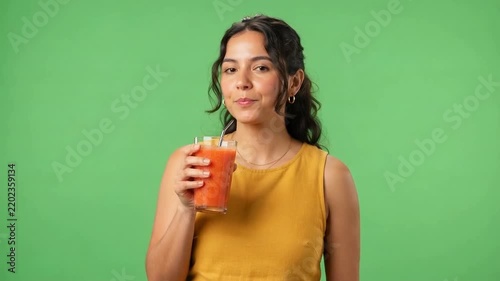 Young woman in a mustard sleeveless top drinks orange juice through a straw against a bright green studio background, smiling with a relaxed expression