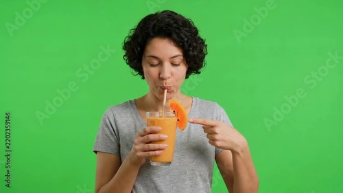 Young woman with curly hair drinks an orange beverage through a straw against a bright green chroma key background