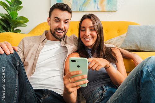 A cheerful pair lounges close together on a bright sofa, looking at a handheld device and sharing a lighthearted moment. The scene conveys connection, curiosity, and vibrant digital interaction