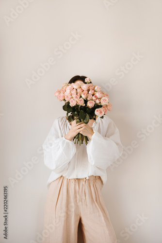Young woman covering eyes with fresh pink rose flowers bouquet in neutral outfit. Aesthetic artistic portrait