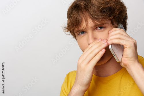 Suspicious young man in a yellow t-shirt covers his lower face while talking on a smartphone, listening to a questionable phone call with a wary, doubtful expression. Concept of scam and distrust.