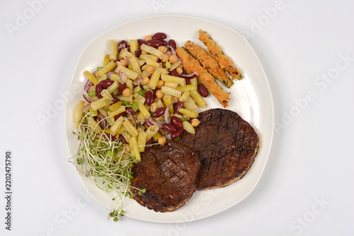 A top down view of a white plate with scored grilled celeriac steaks served alongside bean salad breaded celery sticks and fresh sprouts on a white background.