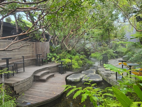 A lush tropical garden cafe with curved wooden decks, black tables, and a misty stream crossed by stepping stones amid dense greenery.