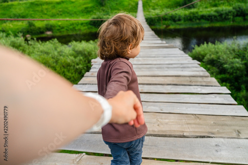 A father holds his sons hand while guiding the little boy across a wooden bridge, ensuring a safe family walk. The child feels secure and trusting as they step together through the greenery.