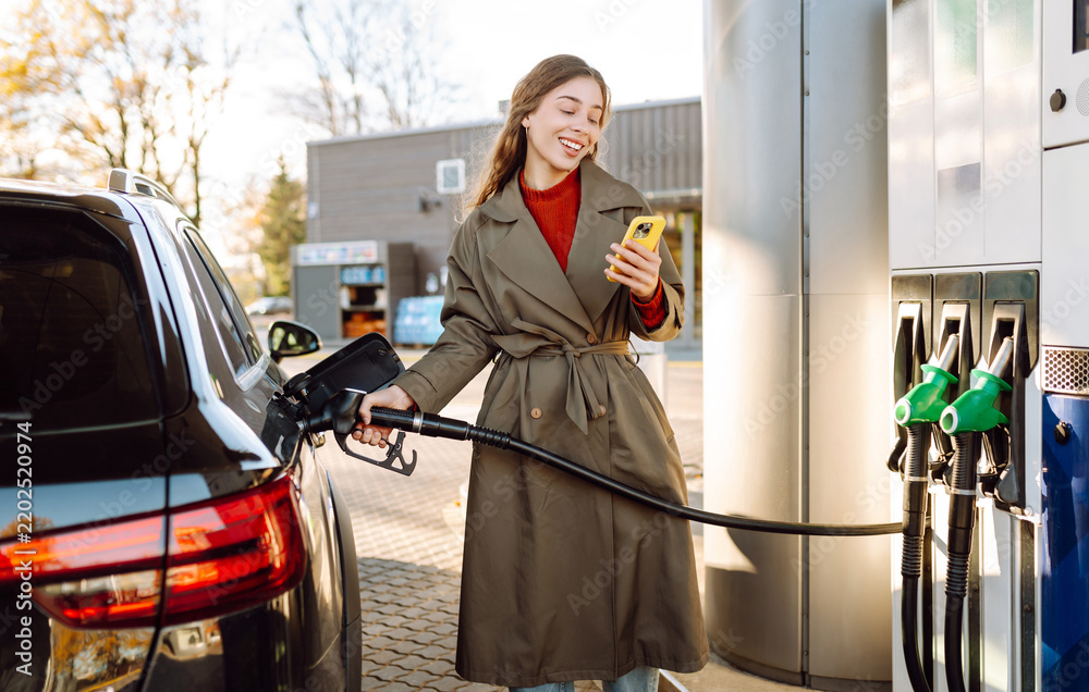 Obraz premium Young woman stands at gas station with phone in hand, refueling her car. Woman with a smartphone at a gas station at sunset. Concept of transportation, refueling, and technology.