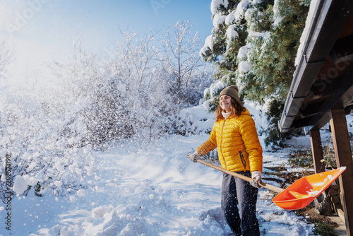 Happy caucasian woman smiling and shoveling snow near a country house terrace on a sunny winter day.