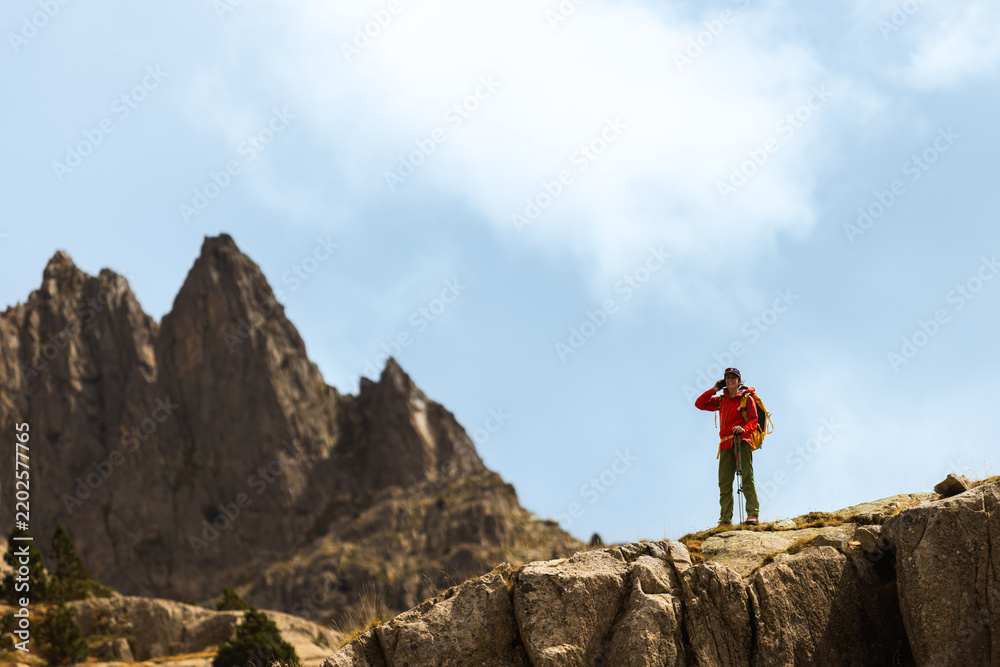 Fototapeta premium Hiker stands on a rocky cliff edge. They wear a red jacket and carry a backpack