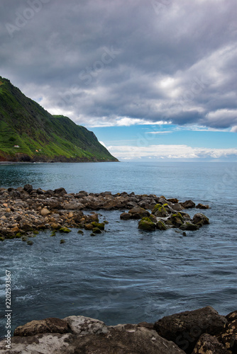Povoação coastline showcasing volcanic rocks in the ocean foreground, leading to calm blue waters and lush green cliffs under a cloudy sky in São Miguel, Azores