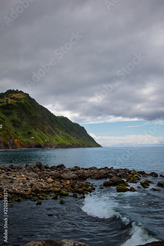 Atlantic Ocean waves crashing against the rugged Povoação coastline, revealing volcanic rocks and vibrant green hills under a cloudy sky in São Miguel, Azores