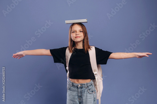 Young girl balancing a book on her head with arms outstretched, wearing a black t-shirt and backpack against a solid blue background