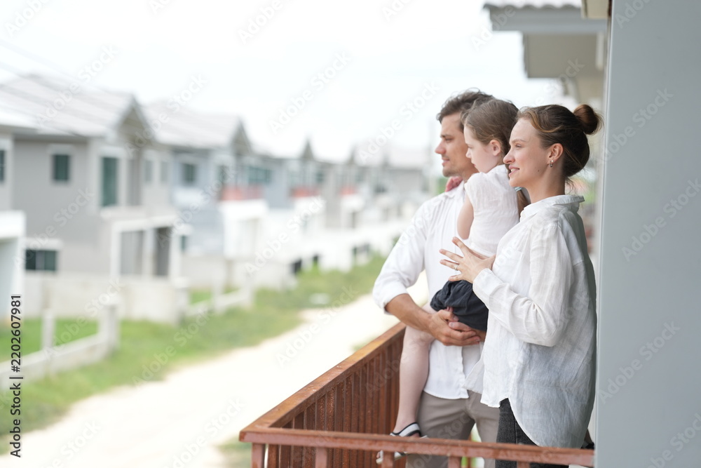 Obraz premium Young parents holding their child look out over the rows of new white houses from the railing on the balcony of their new house overlooking the residential estate, authentic everyday scene
