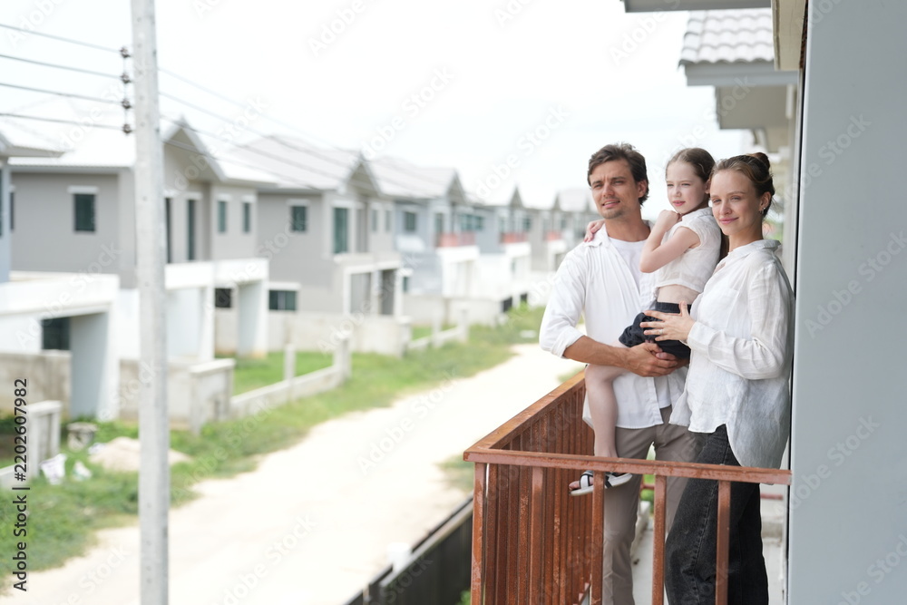Obraz premium Caucasian family of three embracing look out over the rows of new white houses from the railing on the balcony of their new house overlooking the residential estate on a bright sunny day