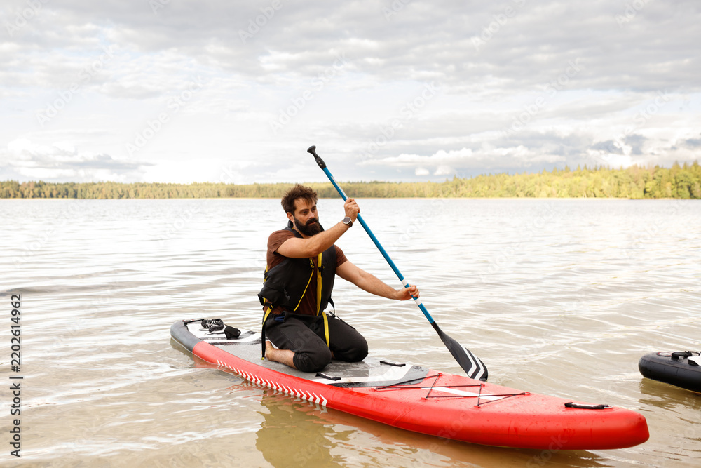 Obraz premium Three quarter front view of a bearded man kneeling and paddling with a blue shaft paddle on a red inflatable SUP board, calm lake water, distant green forest under cloudy sky, daytime horizontal shot