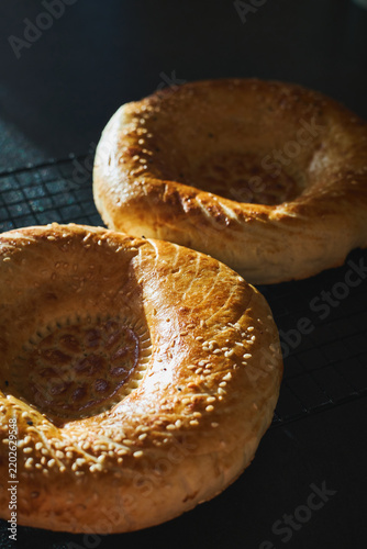 Two golden Uzbek non bread loaves with decorative center pattern, sprinkled with sesame seeds, cooling on metal rack. Homemade baking, traditional Central Asian flatbread.