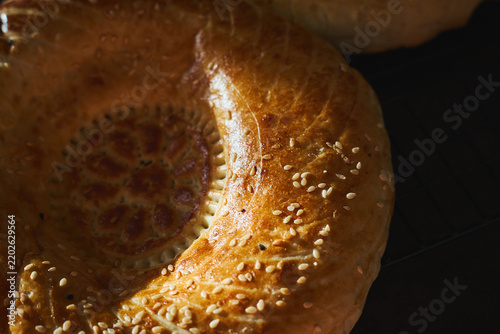 Two golden Uzbek non bread loaves with decorative center pattern, sprinkled with sesame seeds, cooling on metal rack. Homemade baking, traditional Central Asian flatbread.