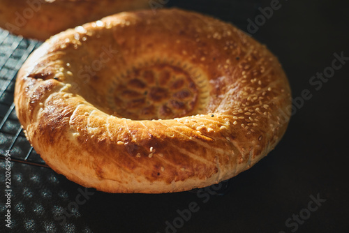 Two golden Uzbek non bread loaves with decorative center pattern, sprinkled with sesame seeds, cooling on metal rack. Homemade baking, traditional Central Asian flatbread.