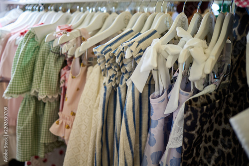 Assorted children's clothing in various colors and patterns including floral, gingham, and stripes hanging on racks and in closets, featuring cotton, linen and muslin fabrics in boutique or home setti