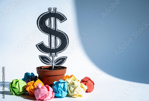 Dollar sign plant in pot with colorful crumpled paper balls on white surface under bright light financial growth