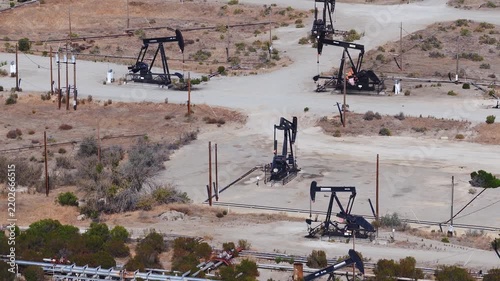 Aerial view showing multiple mechanical pumpjacks extracting petroleum across an arid oil field in California, connected by utility lines and pipelines. Oil tariffs, trade war, price plunge.