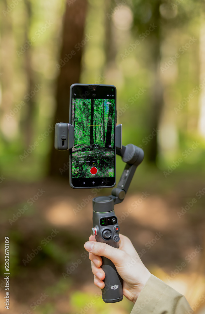 Obraz premium Smartphone video shooting process with professional stabilizer in park. Cameraman hands hold modern telephone device on gimbal setup.
