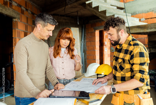 Adult couple reviewing house blueprints with contractor on construction site.