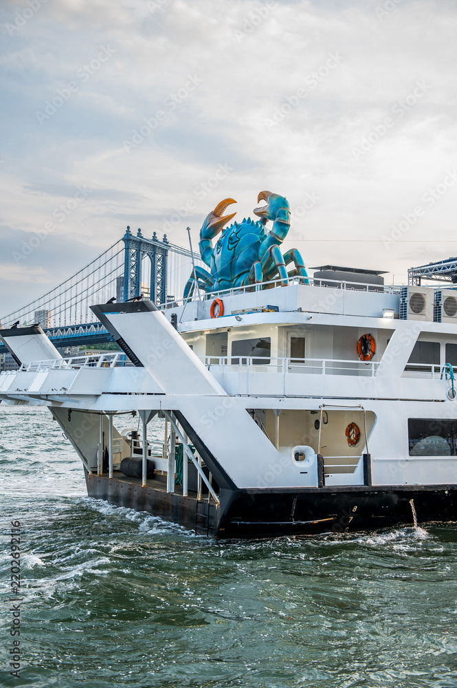 Fototapeta premium Moored vessel with a blue crab sculpture and bridge behind. A crab sculpture sits on a white vessel in New York City, with a steel suspension bridge in the background.