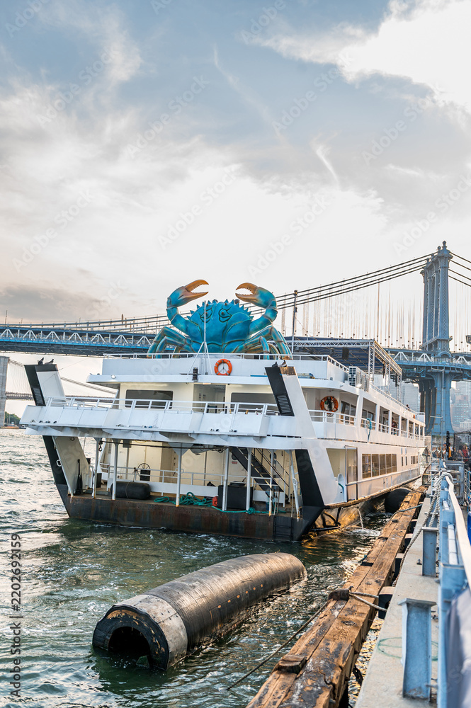 Fototapeta premium Crab sculpture above a moored vessel and floating fender. A white boat sits beside a pier in New York City, with a black fender in the water and a bridge behind.